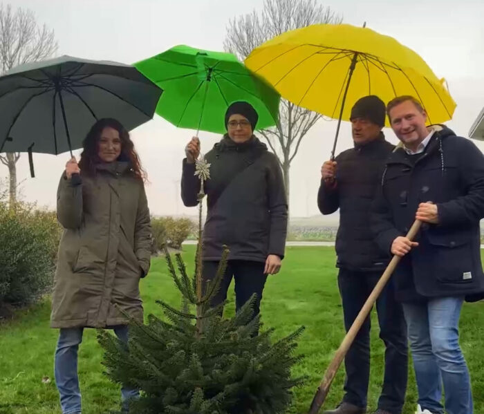 Vier Personen stehen im Freien auf einer Wiese mit einem kleinen Tannenbaum. Zwei halten Regenschirme in Grau, Grün und Gelb. Eine Person hält eine Schaufel. Alle tragen wetterfeste Kleidung. Der Hintergrund zeigt Bäume und einen grauen Himmel.